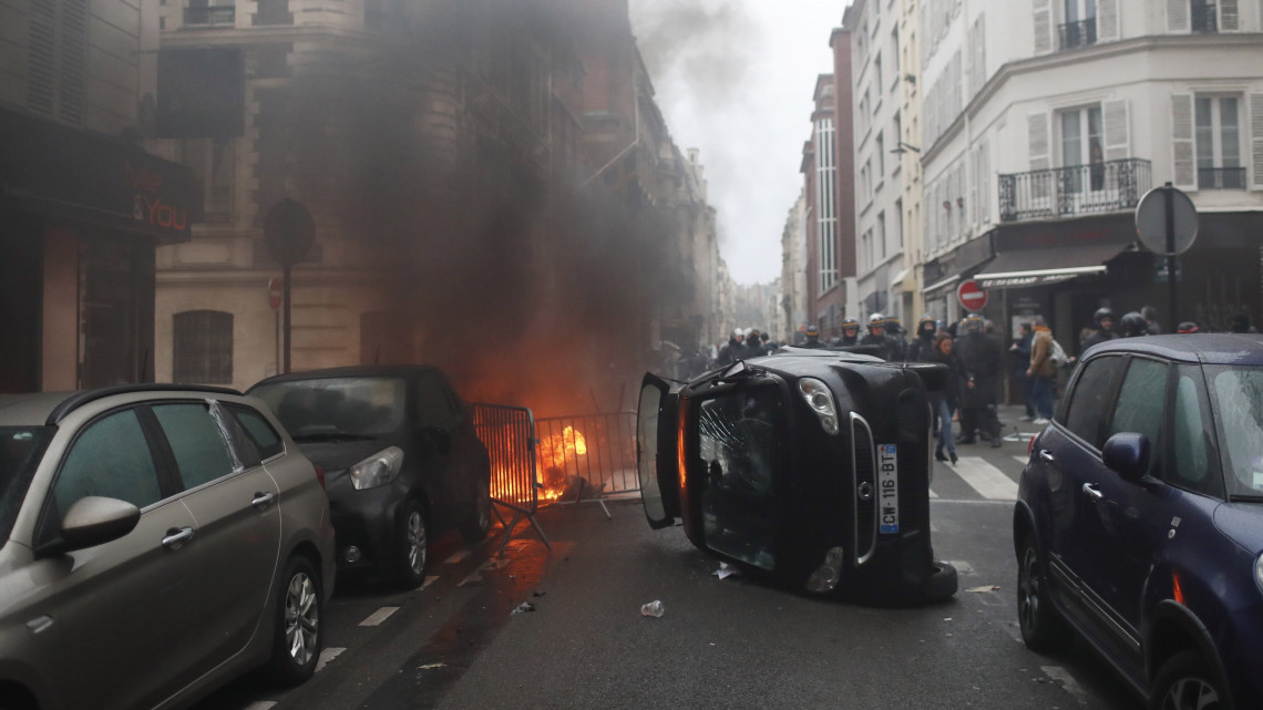 Felgyújtott szemeteskuka lángol egy oldalára döntött autó mögött a francia kormány szociális és adópolitikája ellen tiltakozó tüntetők és a rendőrök összecsapása után Párizsban 2018. december 8-án.