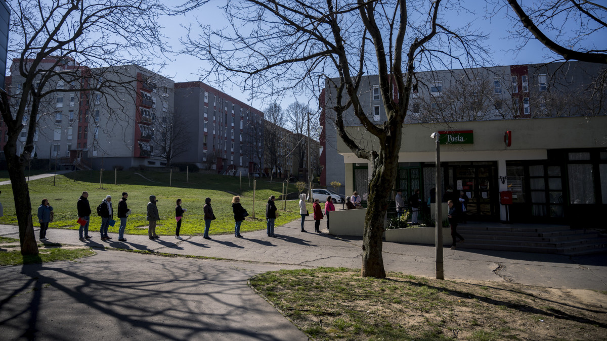 Sorban állók a pécsi 18. számú posta épülete előtt 2020. március 16-án. Az új koronavírus-járvány terjedésének megfékezése érdekében csak annyi ügyfél tartózkodhat a postán, ahány ablak nyitva van.