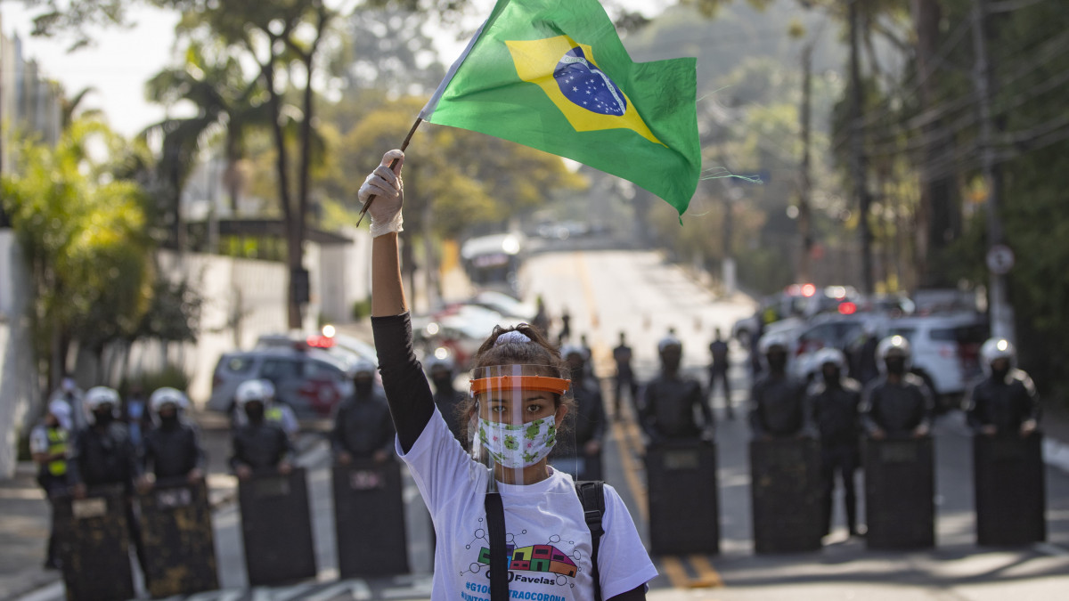 Arcpajzsot és védőmaszkot viselő tüntető egy brazil lászlót lenget a rohamrendőrök sorfala előtt Paraisopolisban, Sao Paulo legnagyobb nyomornegyedében 2020. május 16-án. A tiltakozók azt követelik, hogy a város kormányzója tegye meg a megfelelő óvintézkedéseket a koronavírus-járvány ellen a nyomornegyedekben is.