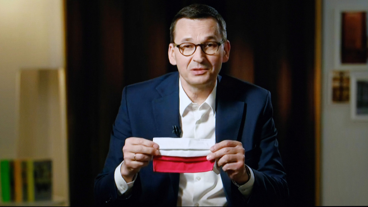 Mateusz Morawiecki lengyel miniszterelnök nemzeti színű szájmaszkot fog a kezében egy kijelzőről készített képen a koronavírus-járványról tartott  internetes beszélgetés során Varsóban 2020. április 21-én. A kormányfőt a koronavírus-járványról és a május 10-re kiírt elnökválasztásról kérdezték.