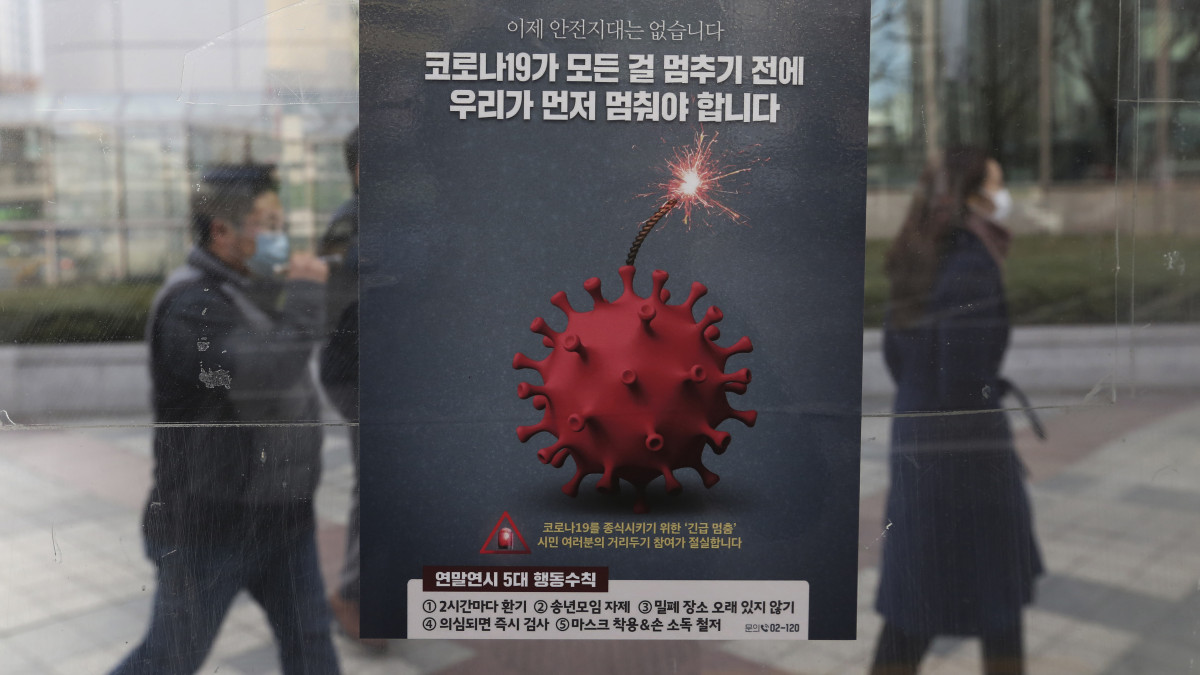 A koronavírus-járvány miatt védőmaszkot viselő emberek haladnak el egy koronavírust ábrázoló poszter előtt Szöulban 2020. december 7-én. A dél-koreai egészségügyi miniszter tájékoztatása szerint Dél-Korea fővárosában és környékén gyorsult a koronavírus-fertőzés terjedése.