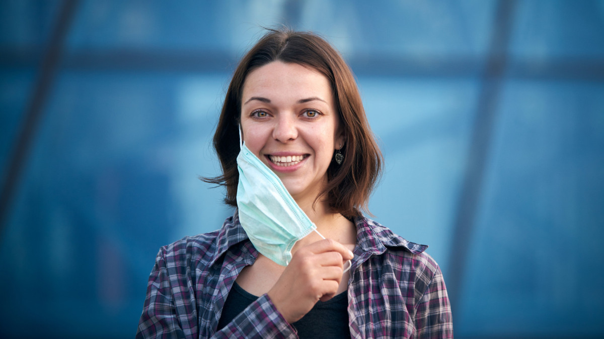 Young happy woman takes off protective medical mask outdoors against modern city background. Pandemic Covid-19 is over concept.