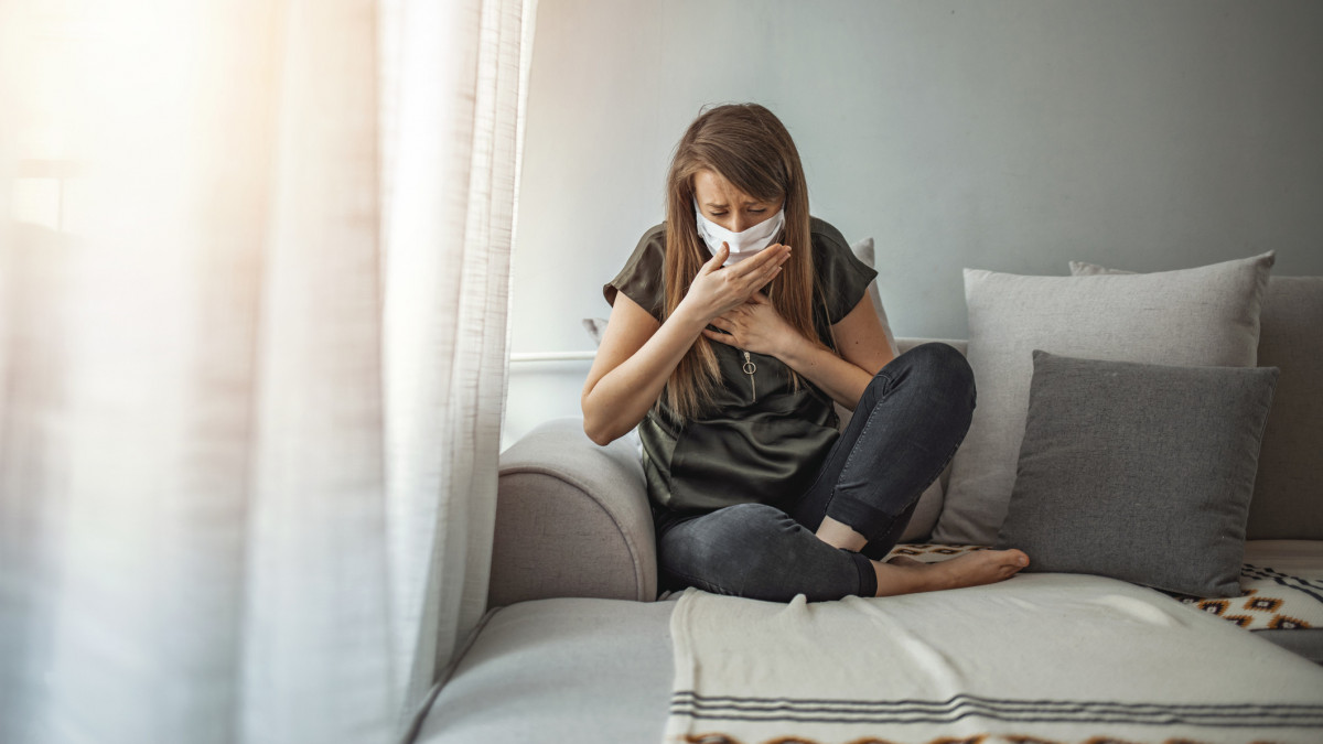 Coronavirus. Woman wearing surgical mask on face protective for spreading of disease Covid-19 pandemic.. Girl symptom cough while sitting on Sofa. Sick woman in face protection mask