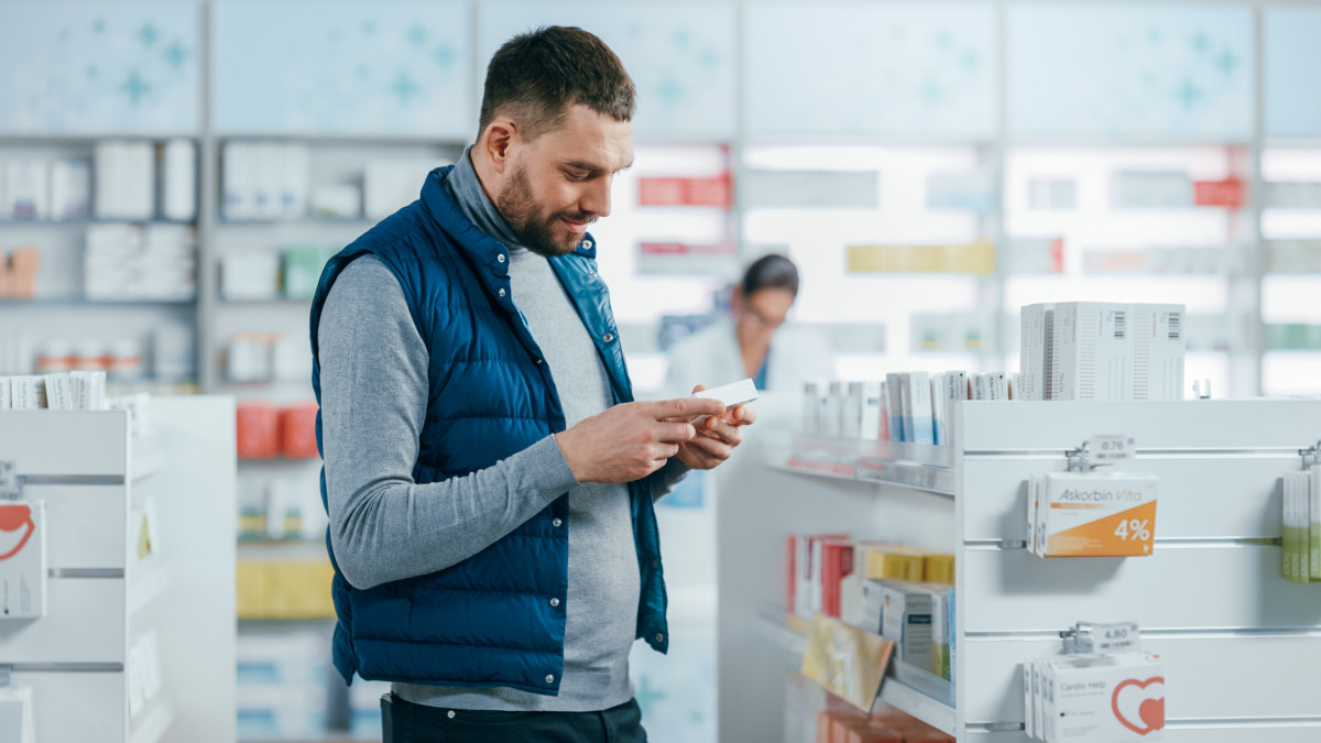 Pharmacy Drugstore: Portrait of a Handsome Young Man Choosing to Buy Medicine, Drugs, Vitamins. Apothecary Full of Health Care Products, Pill Bottles, Sport Supplement Packages with Modern Design