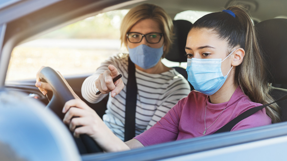 A teenage girl with protective mask sitting behind the steering wheel of a car and listening to her mothers instructions as she drives.