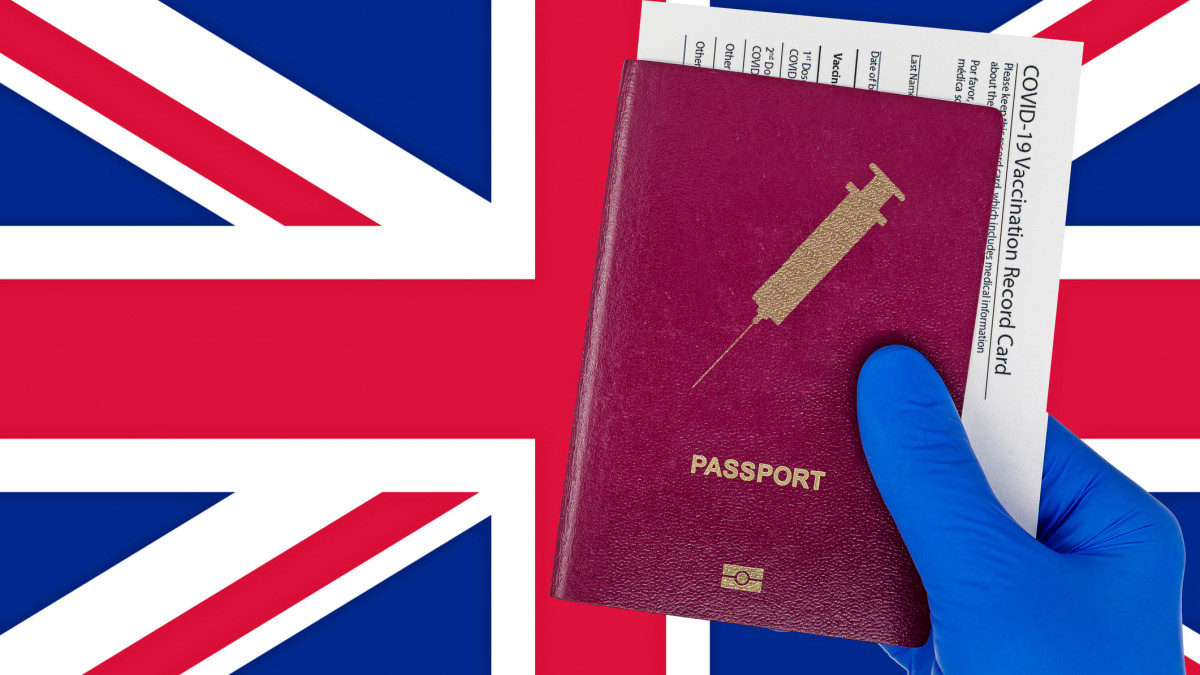 Hand with surgical glove holding a passport with syringe symbol and a vaccination record card or immunization certificate. UK flag on background