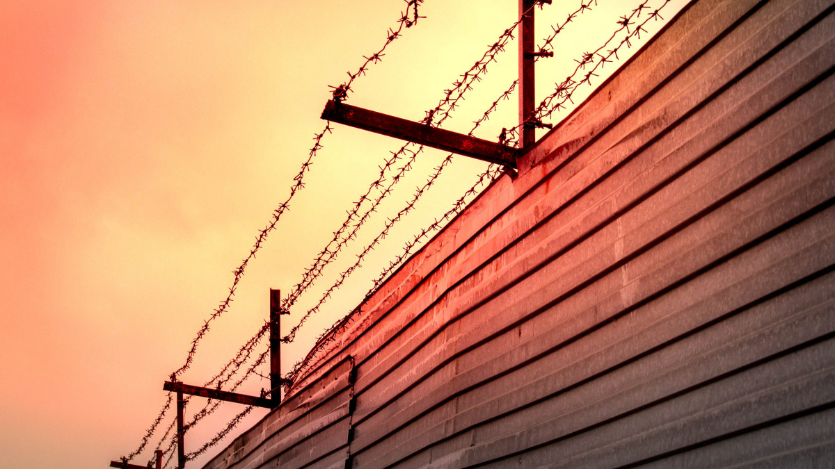 Barbed wire stretched over rusty brackets mounted on a fence made of metal sheets against the backdrop of the red sunset sky.