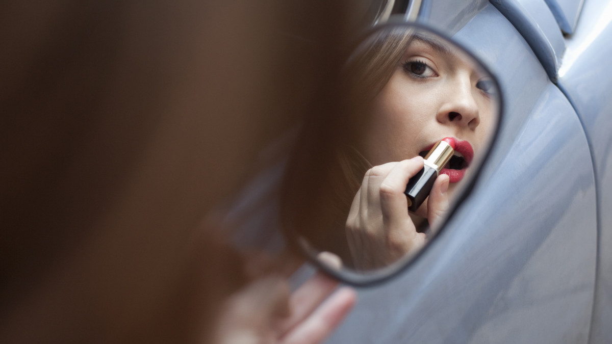 young woman applying lipstick, using rear view mirror of a car