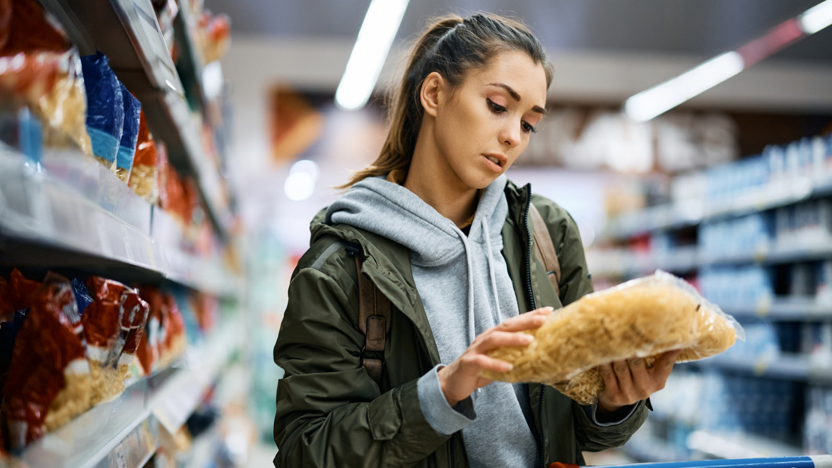 Young woman reading nutrition label while buying pasta at supermarket.