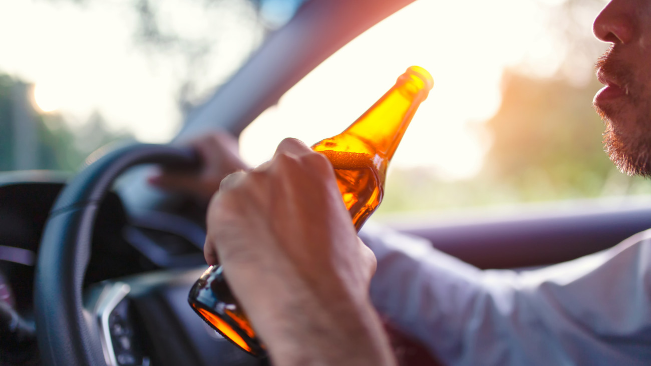 Drunk asian young man drives a car with a bottle of beer with sunset background, Dangerous driving concept