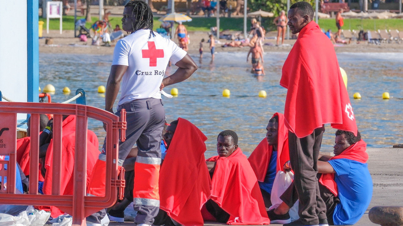 Afrikából Európába igyekvő illegális bevándorlók a Kanári-szigetekhez tartozó Tenerifén fekvő Los Cristianos kikötőjében, a háttérben strandolók 2023. október 26-án. A spanyol parti őrség hajója 23 migránst vett fedélzetére egy csónakból a Kanári-szigetek partjai előtt.