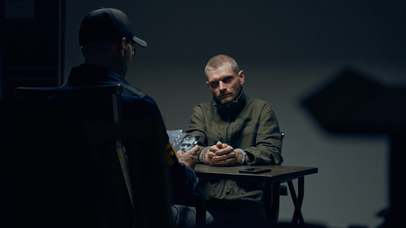 A prisoner in a dark interrogation room sitting in front of an officer with photos of the suspect , we see them sitting at a table. Action movie scene with an officer and a suspect in a dark interrogation room