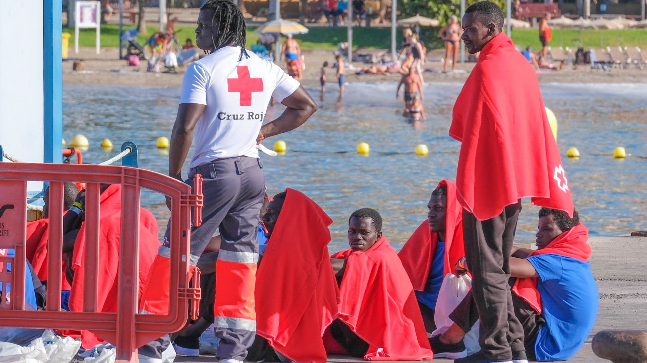 Afrikából Európába igyekvő illegális bevándorlók a Kanári-szigetekhez tartozó Tenerifén fekvő Los Cristianos kikötőjében, a háttérben strandolók 2023. október 26-án. A spanyol parti őrség hajója 23 migránst vett fedélzetére egy csónakból a Kanári-szigetek partjai előtt.