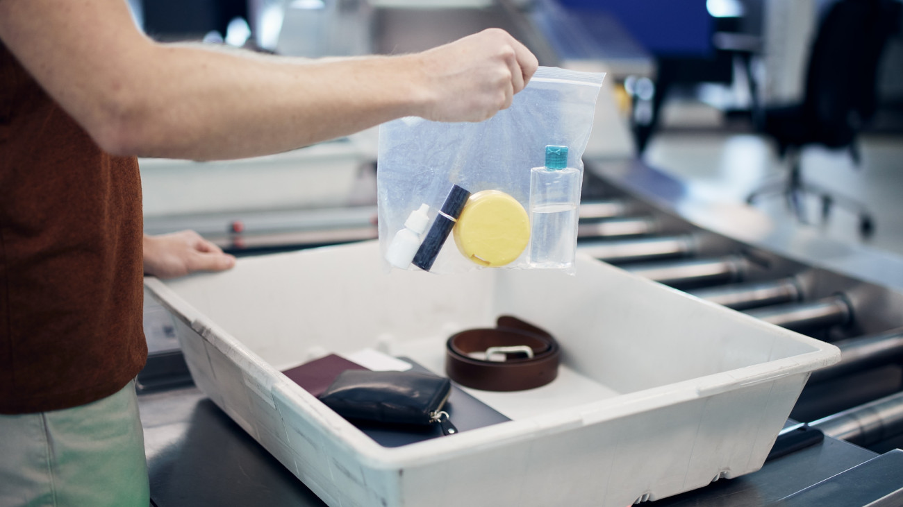 Airport security check before flight. Passenger holding plastic bag with liquids above container with laptop and personal items.