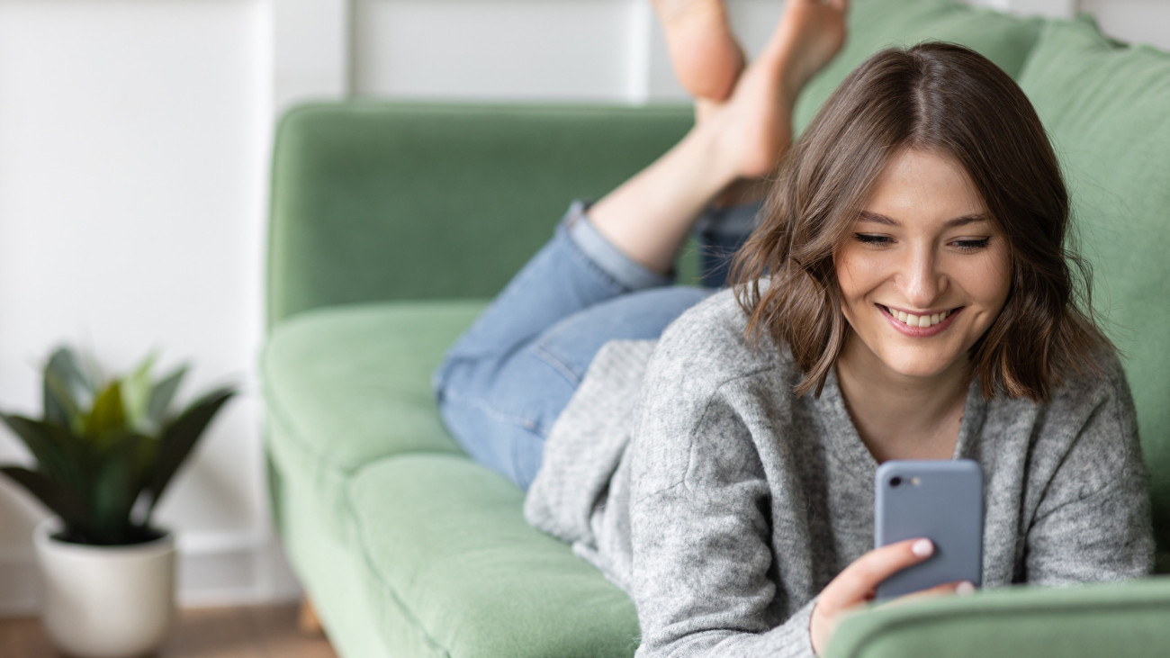 Young beautiful smiling woman using smart phone lying on the couch. Concept of freelance job, home office. Female using technology for learning, shopping, e-commerce, communication, social media