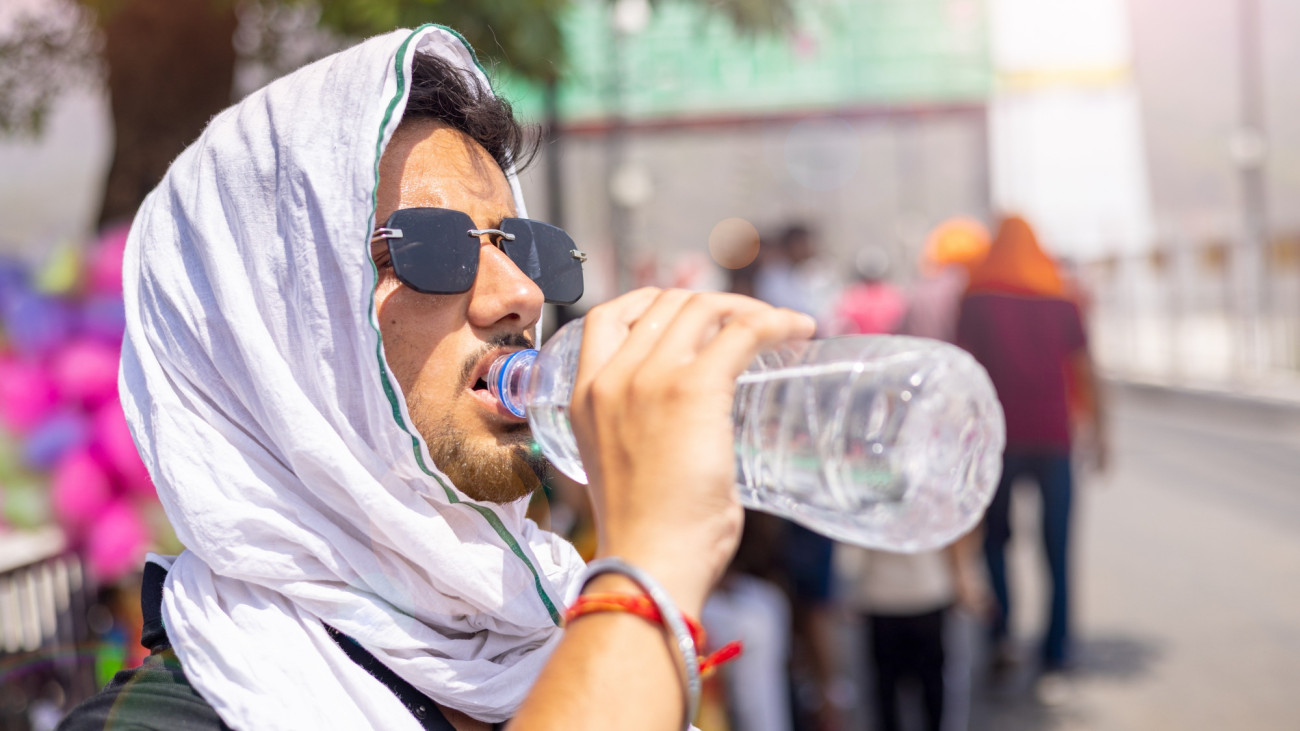 Close-up of a young man covering his head with a cloth and wearing black sunglasses on an extremely hot afternoon while drinking water from a plastic bottle.