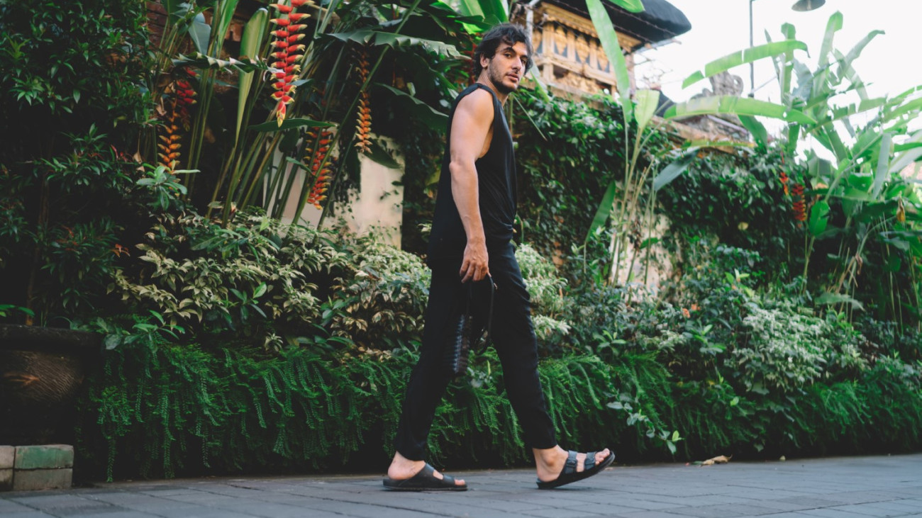 Ground level of ethnic male tourist with bag looking at camera and strolling in tropical park while resting on summer vacation in Indonesia