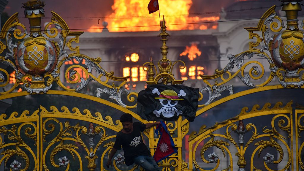KATHMANDU, NEPAL-SEPTEMBER 9: A man is hanging a pirate flag as smoke and flames rise from the Singha Durbar after people set fire to the Singha Durbar, the seat of Nepal governments various ministers offices in Kathmandu, Nepal on September 9, 2025. At Least 19 people were killed and dozens injured on September 8 during the demonstration against corruption and ban on social media by the government. (Photo by Sunil Pradhan/Anadolu via Getty Images)