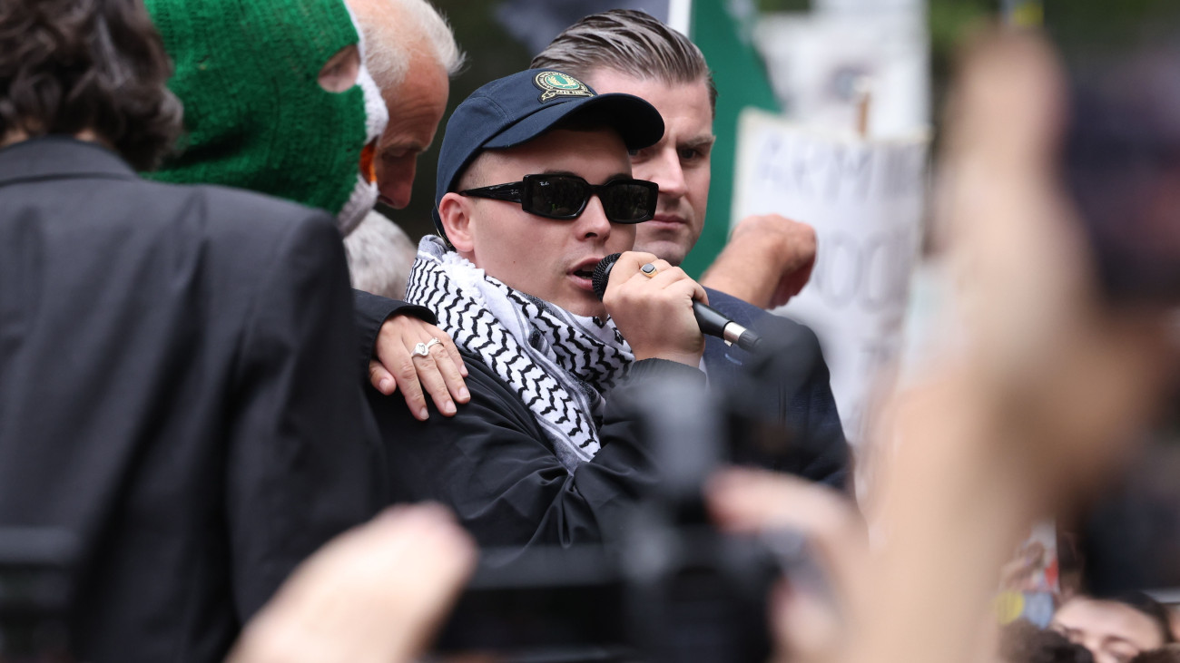 Liam Óg Ó hAnnaidh, művésznevén Mo Chara, a Kneecap északír rapegyüttes egyik tagja beszél támogatóihoz a londoni Westminster kerületi bíróság előtt 2025. augusztus 20-án, a terrorizmus bűncselekményének gyanúja miatti kihallgatásuk után. A belfasti zenészek az áprilisi kaliforniai Coachella fesztiválon rendezett fellépésükön palesztinpárti és Izrael-ellenes üzeneteket jelenítettek meg, valamint az egyik 2024 novemberi fellépésükön a Hezbollah libanoni síita szervezet zászlaját lengették.