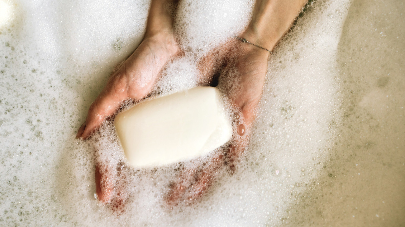 White toilet soap in female hands against the background of a fragrant foam bath. Woman in white terry bathrobe washing her hands with soap. Relaxation and beauty treatments. Natural beauty, daily skincare routine. Moisturizing, cleansing