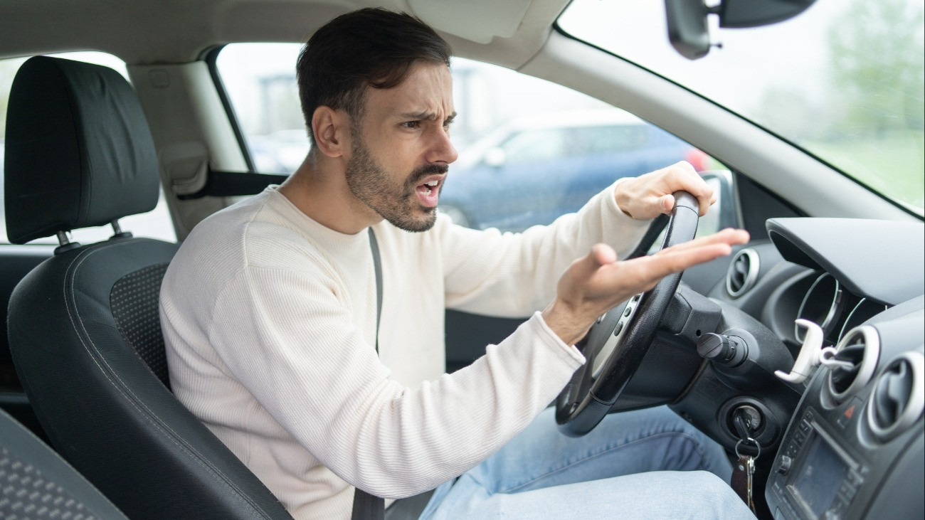 A man in a car shows frustration, gesturing with his hands.