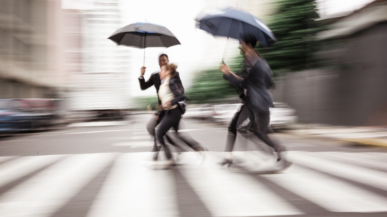 Three people with umbrellas hurry across a crosswalk on a rainy day, creating a sense of urgency and motion amidst urban surroundings.
