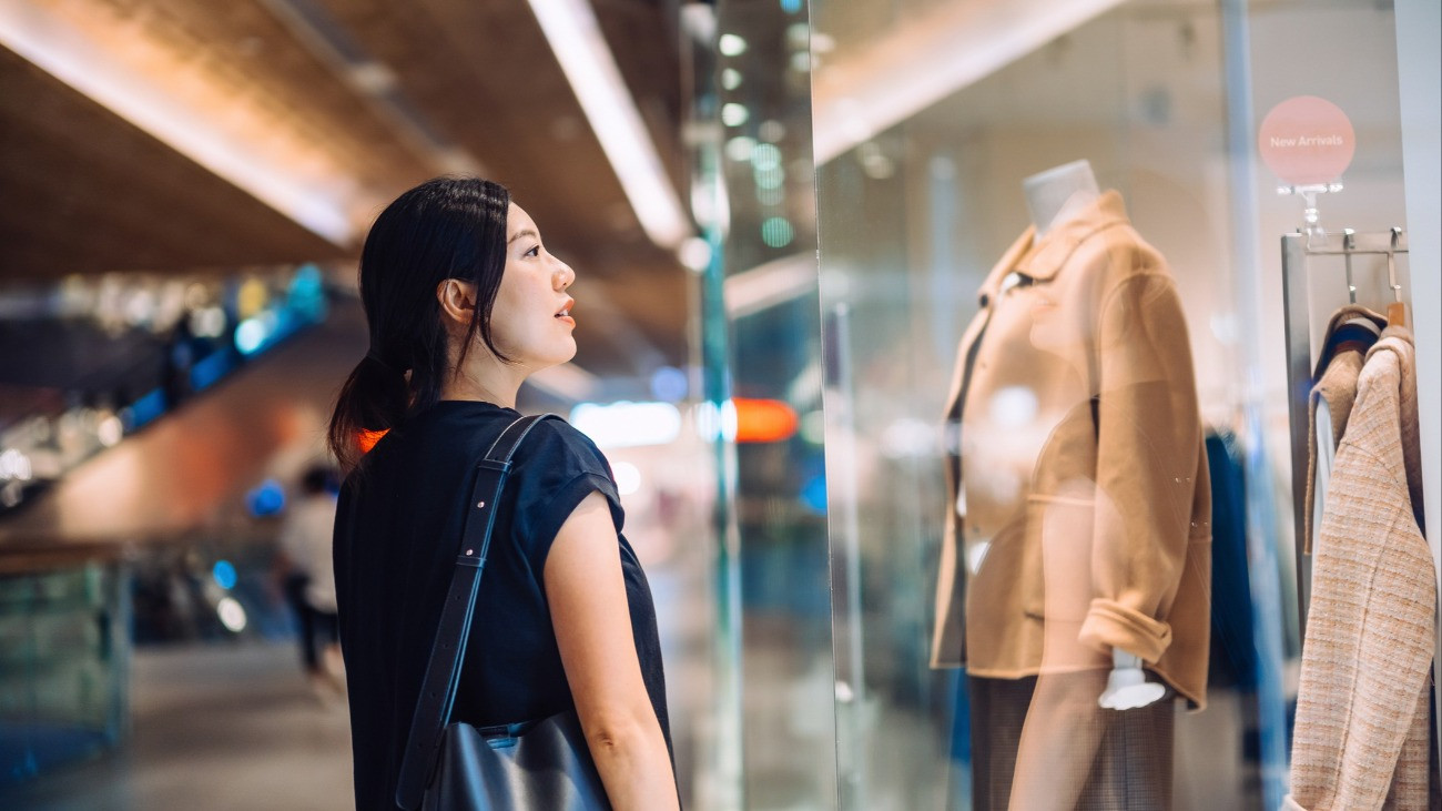 Fiatal ázsiai nő áll egy butik előtt, és a kirakatot nézi vásárlás közben a bevásárlóközpontban.