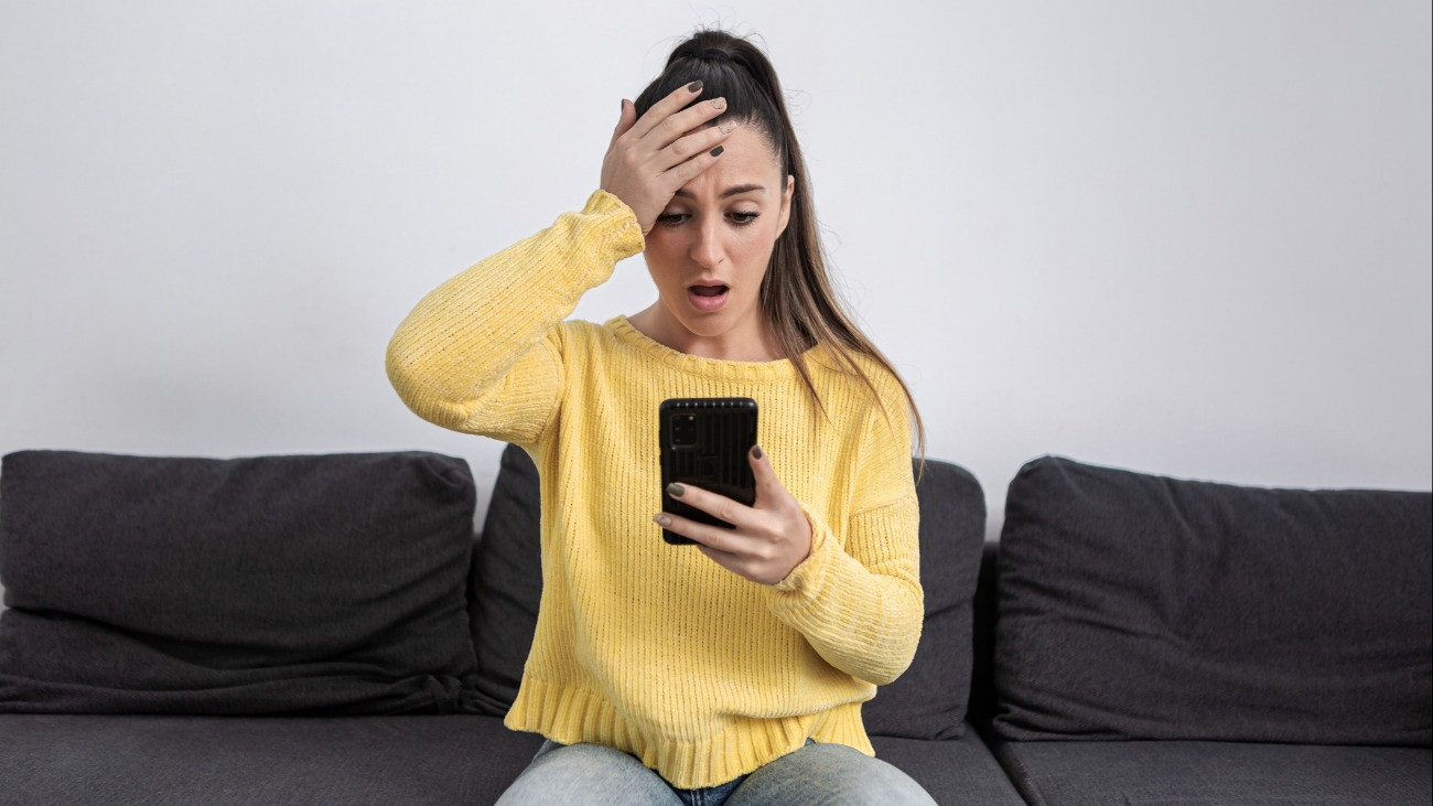 Young amazed female touching head and opening mouth in fear while looking at smartphone screen getting scammed online