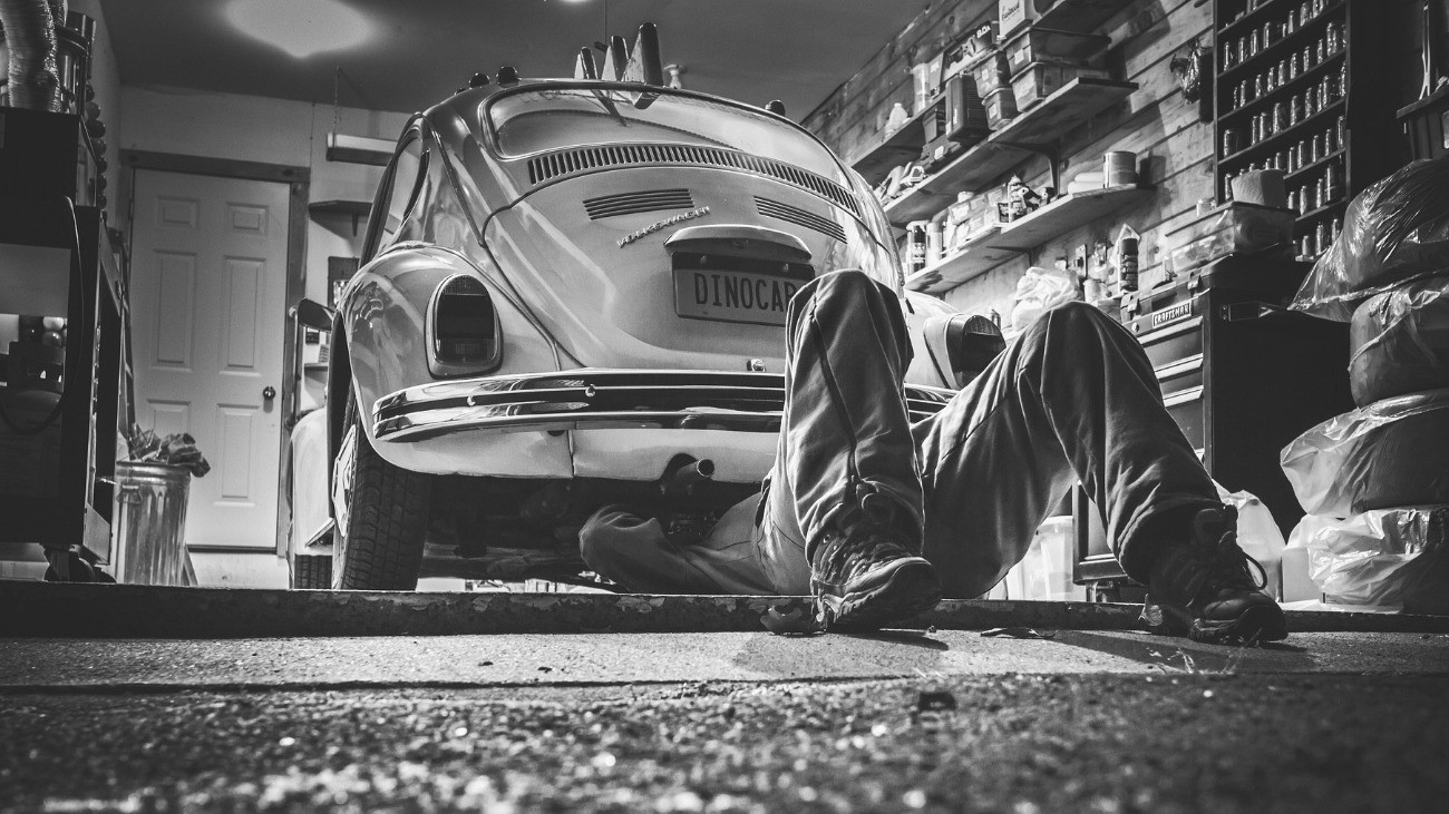 Fekete-fehér fotón egy műhely látszik, ahol egy bogárhátú Volkswagen motorja alatt fekve tevékenykedik a szerelő.