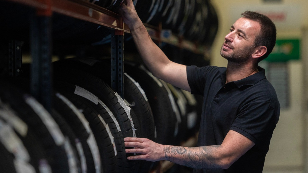 Egy tetovált férfi mosolyogva nézegeti az új gumiabroncsokat egy autószerviz raktárában.