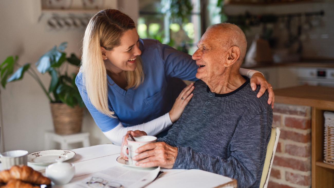 Happy young female caregiver or healthcare worker visiting senior man indoors at home, talking and smiling.