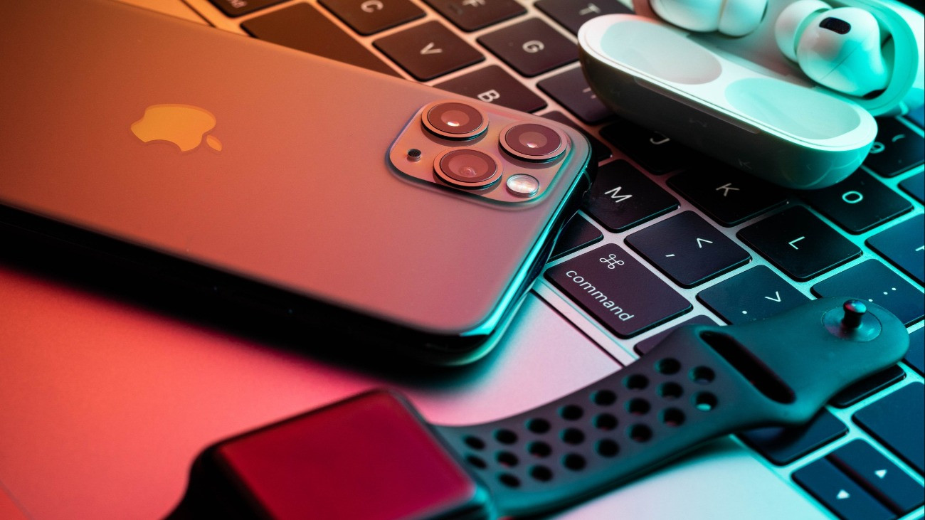 iPhone 11 Pro with AirPods Pro and Apple Watch with a Macbook in the background with teal and orange glow.