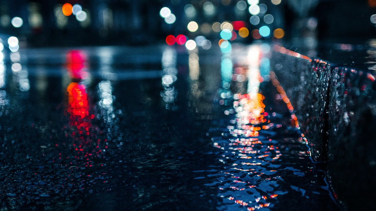 Éjszakai fények esőben, ahogy egy járdán és úttesten visszatükröződnek.