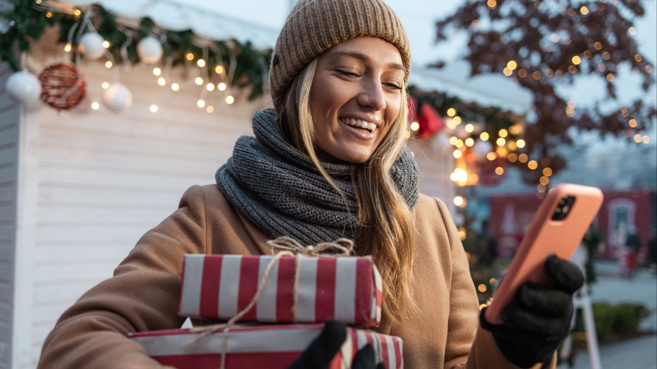 Photo of a happy young woman holding Christmas gifts and looking at her phone, enjoying the festive holiday outdoors. She is smiling and appears excited, embracing the joyful holiday spirit.