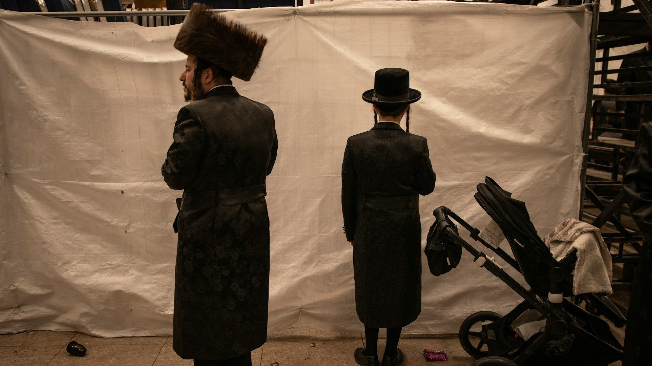 Két ultraortodox zsidó férfi egy babakocsi mellett, egy paravánnal szemben áll, háttal a kamerának.
