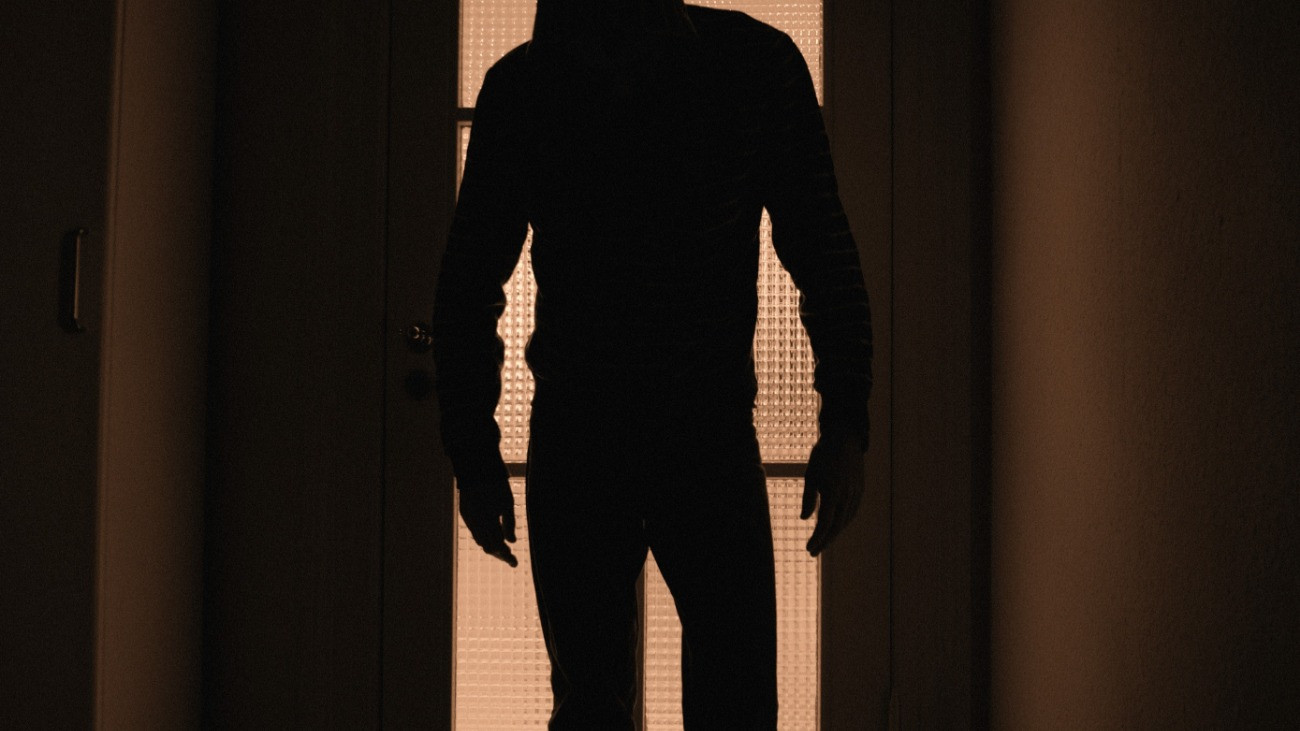 Férfi sziluettje fényes háttér előtt egy ajtóban / Hooded man standing in a somewhat threatening and spooky pose in front of a glass door. The man is standing in a dark room - the light is lit in the room behind the door. Focal point is on the door behind the man. Grainy.