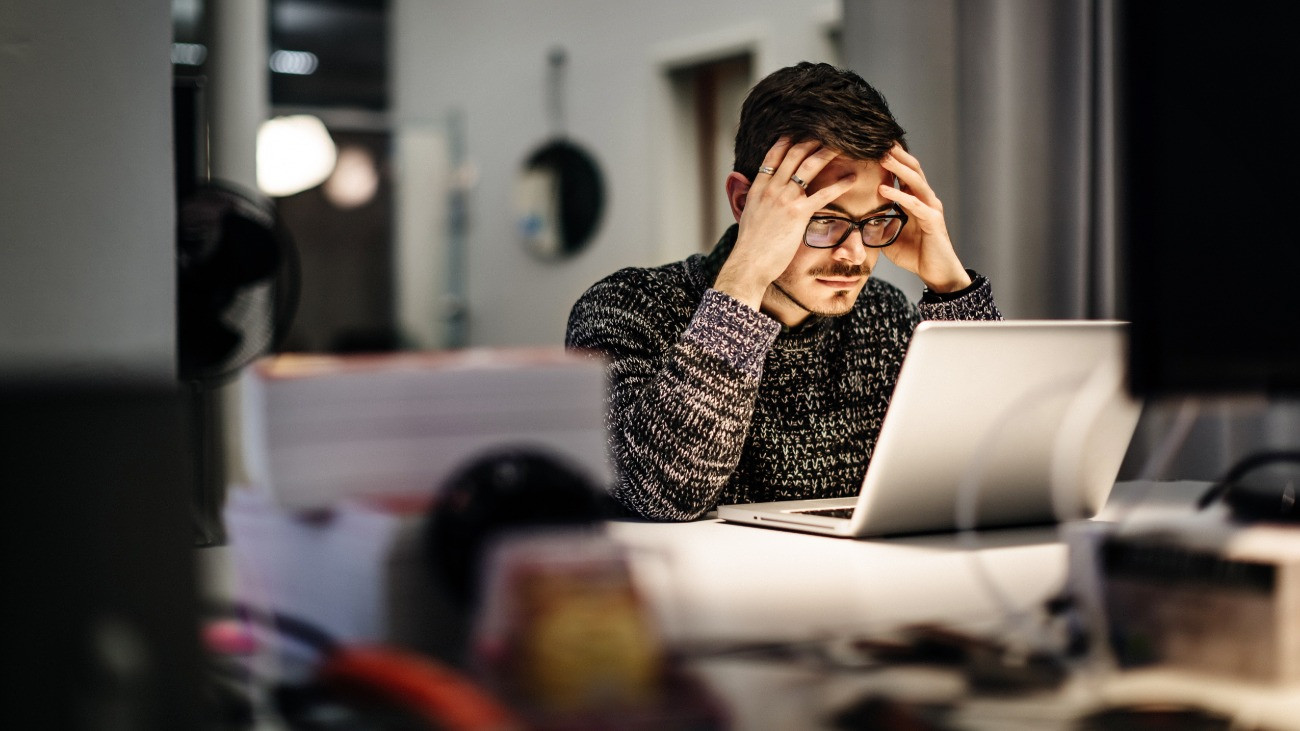 Fiatal üzletember gondterhelten nézi egy laptop kijelzőjét egy irodában // Young casual businessman wearing glasses is sitting in front of his notebook holding his head pondering over his work. Office equipment and another computer is in front of him.