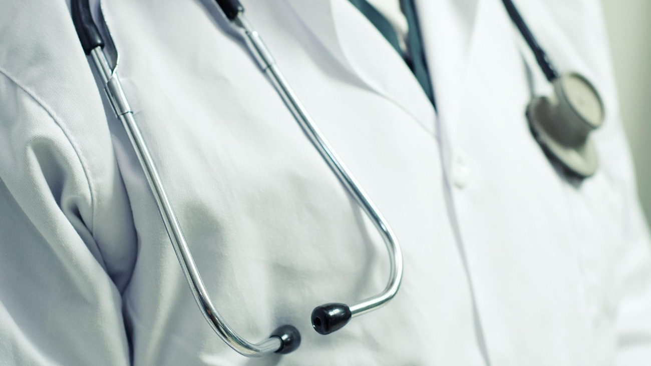 Közeli kép egy fehér köpenyes orvos nyakában elhelyezkedő fonendoszkópról, egy kézi diagnosztikai eszközről, amely például a szív és a tüdő hangját akusztikailag fölerősítve és mindkét fülhöz vezetve segíti az orvos munkáját.