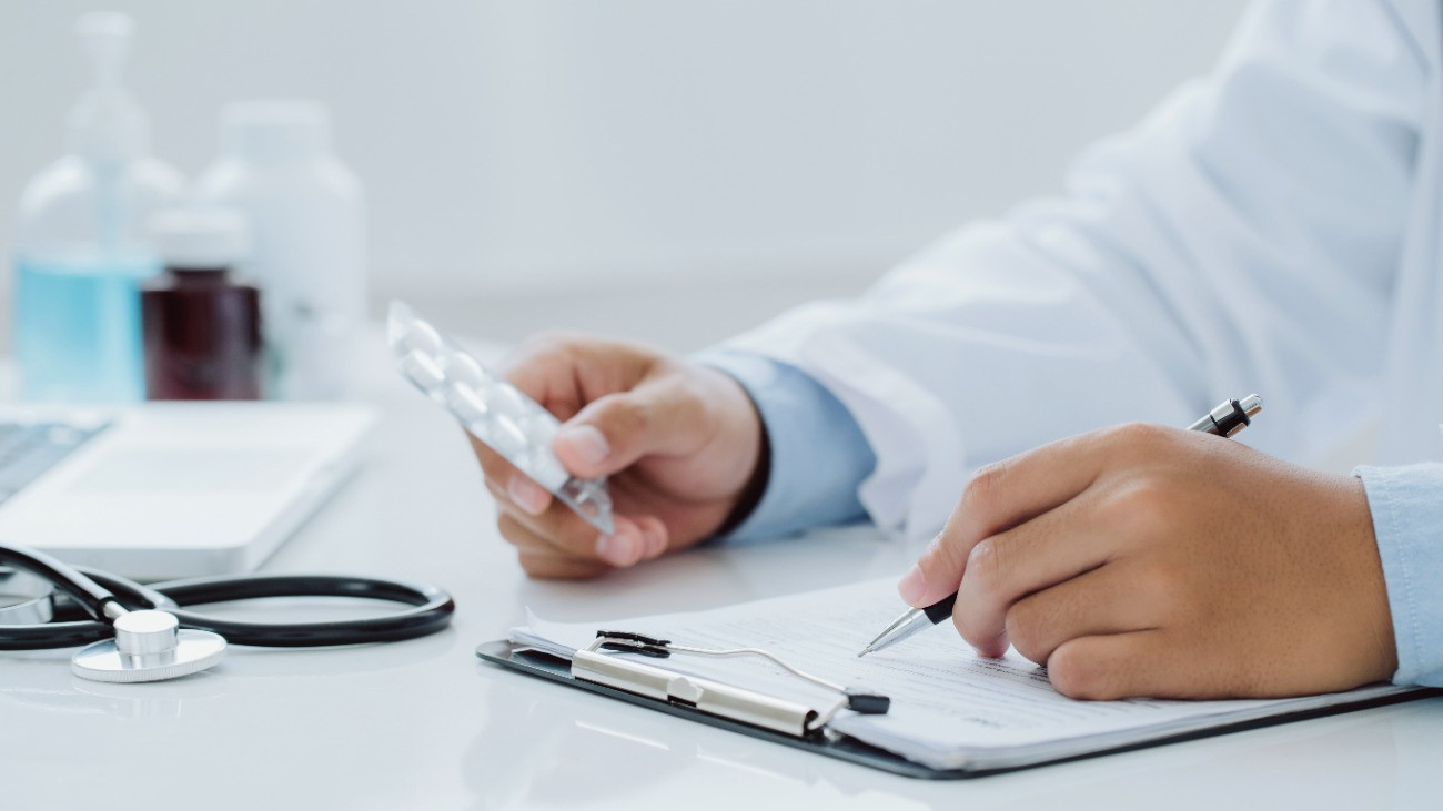Receptet ír fel egy orovos a betegnek, miközben az egyik kezében gyógyszert tart.