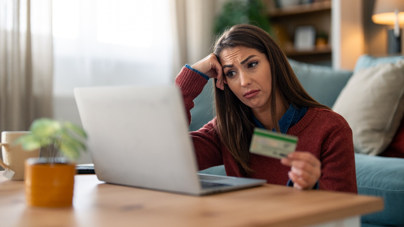 Töprengő tekintetű nő vizslatja a laptopot, kezében egy bankkártyával.