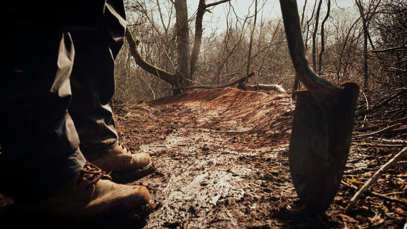 Egy bakancsos alak ásóval a kezében áll egy erdős területen.